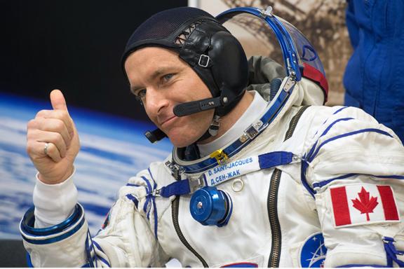 Canadian astronaut David Saint-Jacques is looking to our left, smiling and giving the thumbs-up signal.  He is wearing a Russian cosmonaut Sokol KV2 space suit, without the gloves and with the helmet visor raised.  